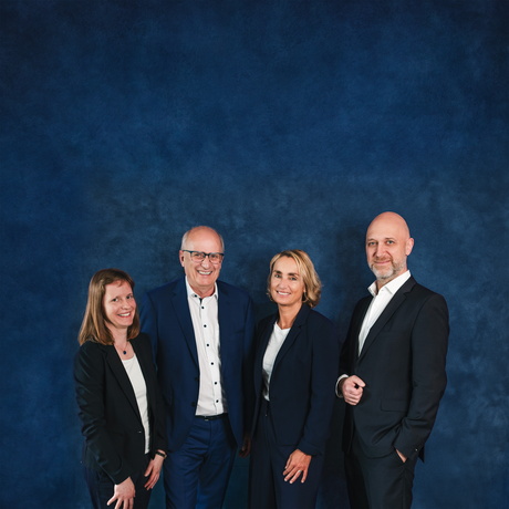 Vier Personen in Businesskleidung stehen nebeneinander vor einem blauen Hintergrund.